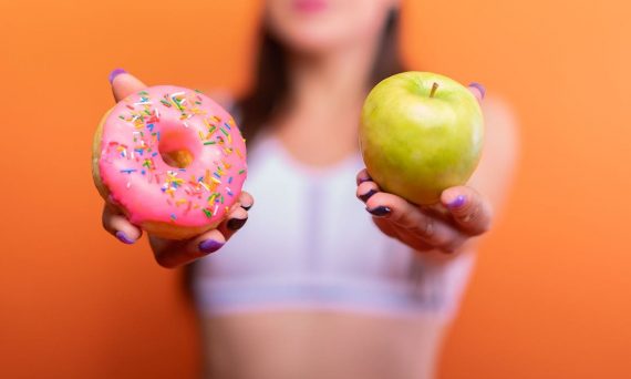A woman holds a green apple and a pink donut with sprinkles against an orange background, emphasizing choice.