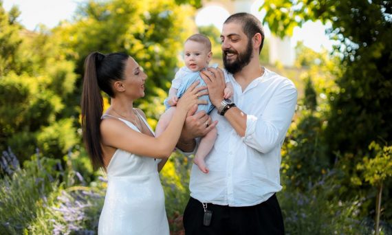 Mother and father joyfully hold their baby in a vibrant outdoor garden setting.